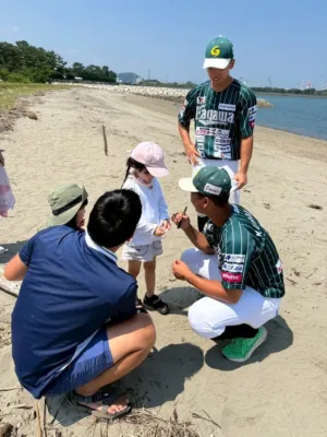晴れた日に砂浜で、野球選手が子供たちと交流している様子です。一人の選手がしゃがんで子供にサインをしており、他の大人や子供たちも見守っています。