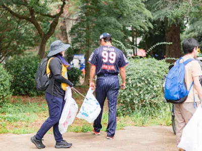 公園でゴミ拾いをするボランティアの人々。野球ユニフォームの男性と帽子をかぶった女性が、火ばさみとゴミ袋を持って清掃活動をしている
