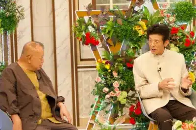 二人の男性が豪華な花の装飾が施されたスタジオで会話している様子です。右側の男性が身振り手振りを交えて話しており、左側の男性は熱心に聞き入っています。