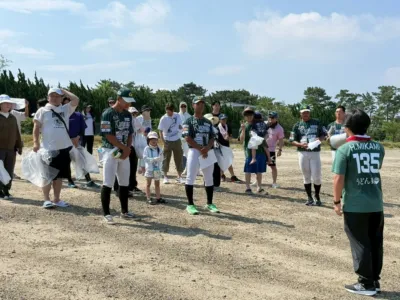 晴れた日に屋外に集まった人々が写っています。野球のユニフォームを着た人たちもおり、清掃活動のような地域イベントに参加しているようです。子供から大人まで多くの人が集まっています。