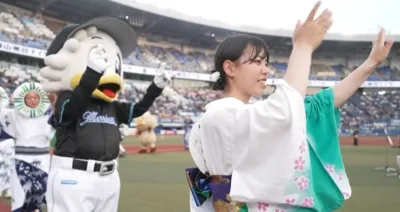 野球場のフィールドで、浴衣姿の女性と野球チームのマスコットキャラクターが観客に向けて手を振っている様子