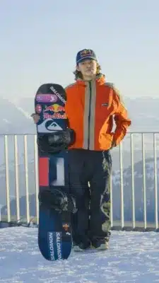雪山でオレンジ色のジャケットを着た男性がスノーボードを抱えて立っています。ボードにはRed BullやSALOMONのロゴが見え、冬のスポーツシーンを捉えた一枚です。