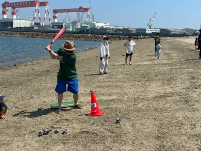 砂浜で大人と子供たちがティーボールのような遊びを楽しんでいる様子。一人の男性がバットを構え、赤いコーンの上に置かれたボールを打とうとしている。背景には海と大きな造船所のクレーンが見える、のどかな夏の日の風景。