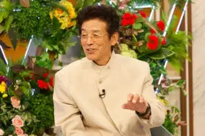 笑顔で話す中高年男性が写っています。ベージュのスーツと眼鏡を着用し、背景には豊かな緑の植物と色とりどりの花が飾られたスタジオのような空間が広がっています。テレビ番組の出演中かインタビューを受けている場面のようです。