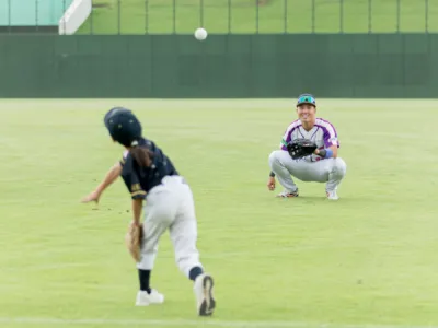 緑の野球場で、子供がボールを投げ、笑顔の大人(選手またはコーチ)が捕球の姿勢で待っている