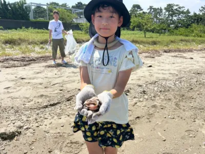 麦わら帽子と手袋を着用した子供が、砂浜で大きな貝殻を両手に持っています。背景には、大人もゴミ袋とトングを持って清掃活動をしている様子が見られ、晴れた日のビーチクリーンイベントのようです。