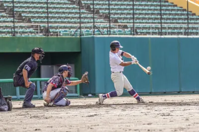 野球の試合で、バッターがボールを打つ瞬間を捉えた写真です。キャッチャーと審判も写っており、緊迫したプレーの様子が伝わります。