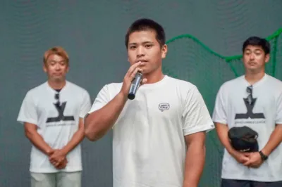 白いTシャツを着た3人の男性が写っており、中央の男性がマイクでスピーチをしています。シャツには「LIBERTY FORCE」や「JAPAN SUMMER LEAGUE」のロゴが見られ、スポーツ関連のイベントや会見の場面を示唆しています。