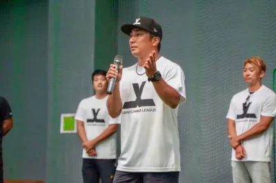 「JAPAN SUMMER LEAGUE」のロゴ入りTシャツを着た男性がマイクを持ってスピーチしています。他の数人の男性も同じTシャツを着用しており、野球関連のイベントや会見の一場面と推測されます。