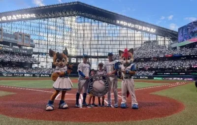 北海道日本ハムファイターズの本拠地エスコンフィールドHOKKAIDOで、マスコット、選手、子供たちがグラウンドに立ち、記念の盾を囲んでポーズをとっている様子
