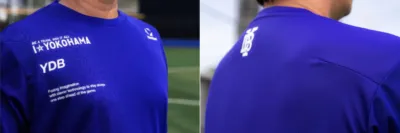 青い長袖シャツを着た人物のクローズアップ画像。シャツには「I☆YOKOHAMA」「YDB」といった文字と、「BE A TEAM, WIN IT ALL」などのスローガンが白字でプリントされています。