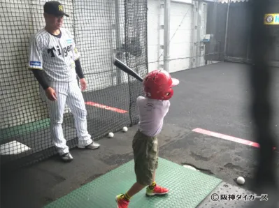 阪神タイガースのユニフォームを着た大人が子供のバッティング練習を見守る様子