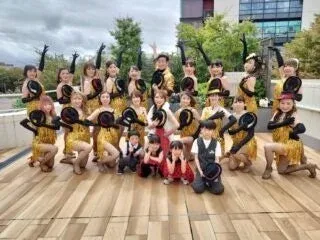 きらびやかな金色のフリンジ衣装をまとった大人数のグループが、屋外の広場で笑顔でポーズをとっている。ダンスパフォーマンス後の記念撮影か、イベントの一場面のようだ。