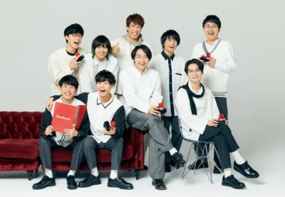 「QuizKnock」のメンバーと思われる9人の男性が、白い背景のスタジオで笑顔でポーズをとっている集合写真です。数名がクイズの早押しボタンを持ち、一人が「QuizKnock」と書かれた赤いファイルを抱えています。明るく和やかな雰囲気が特徴です。