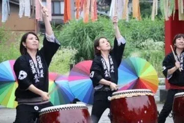 黒い法被を着た女性たちが和太鼓を力強く演奏している様子です。背景には色とりどりの傘が配置されており、お祭りやイベントでの賑やかなパフォーマンスを捉えた一枚です。