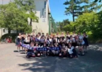 多くの学生が屋外で集合写真を撮っている様子です。皆、楽しそうな笑顔でポーズをとっており、学校の建物と木々を背景に、学校行事かクラブ活動の一コマのように見えます。