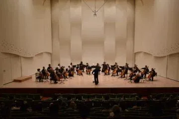 コンサートホールでオーケストラが演奏している様子を捉えた画像です。指揮者が中心に立ち、多くの弦楽器奏者が舞台上で熱心に演奏しており、観客席の一部も映り込んでいます。
