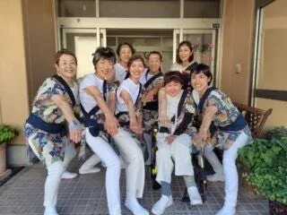 カラフルな衣装を身につけた女性たちのグループが、建物の前で笑顔でポーズをとっている集合写真です。車椅子の方もおり、イベントやパフォーマンスの一幕のようです。