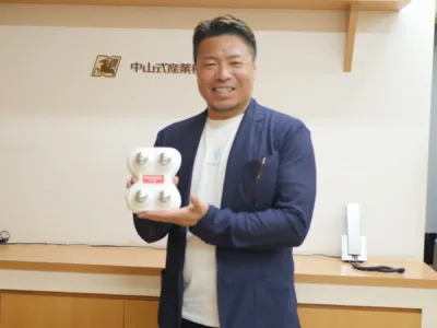 中山式産業のロゴを背景に、ネイビーのジャケットを着た男性が笑顔で白いデバイスを手に持っている
